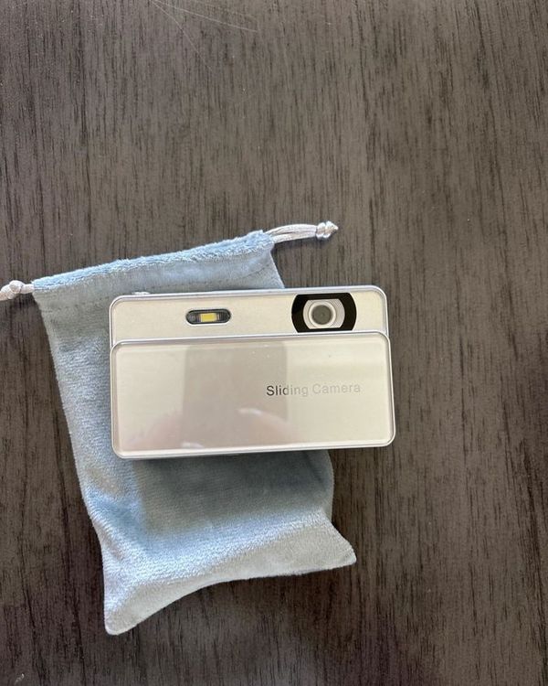 a silver slide camera on a wooden table next to a grey pouch